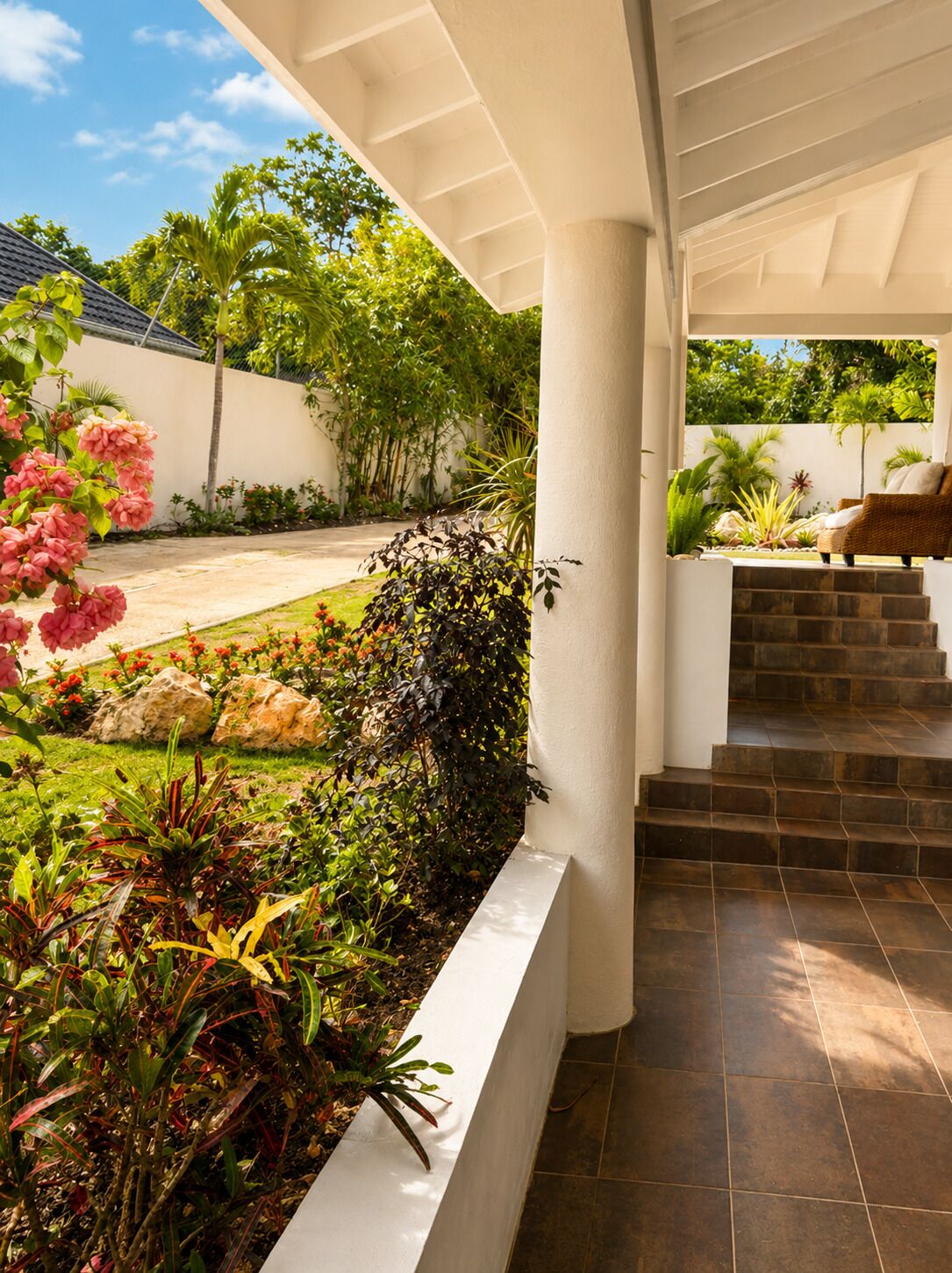 A covered outdoor area with a garden, a white column, and a set of stone steps leading to a seating area.