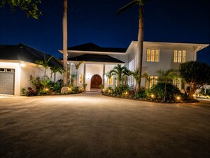 Una casa de dos pisos bien iluminada, con una puerta principal grande, rodeada de palmeras y un camino de entrada que lleva a un garaje.