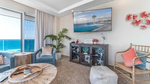 A modern living room with a large window overlooking the ocean, a flat-screen TV mounted on the wall, and a wooden cabinet with decorative items. - Verano 1601 – Spacious Corner Condo (Puerto Peñasco)