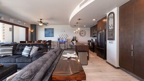 A modern living room with a dark sofa, a wooden coffee table, and a dining area with a curved kitchen in the background. - Las Palomas Phase II Coronado 506 Exceptional Oceanfront 2BR 2BA Unit. (Puerto Peñasco)