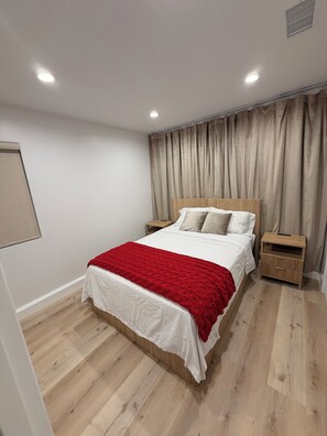 Ein Schlafzimmer mit einem Bett, einem Nachttisch, einer roten Decke, Holzboden und einer Deckenleuchte.