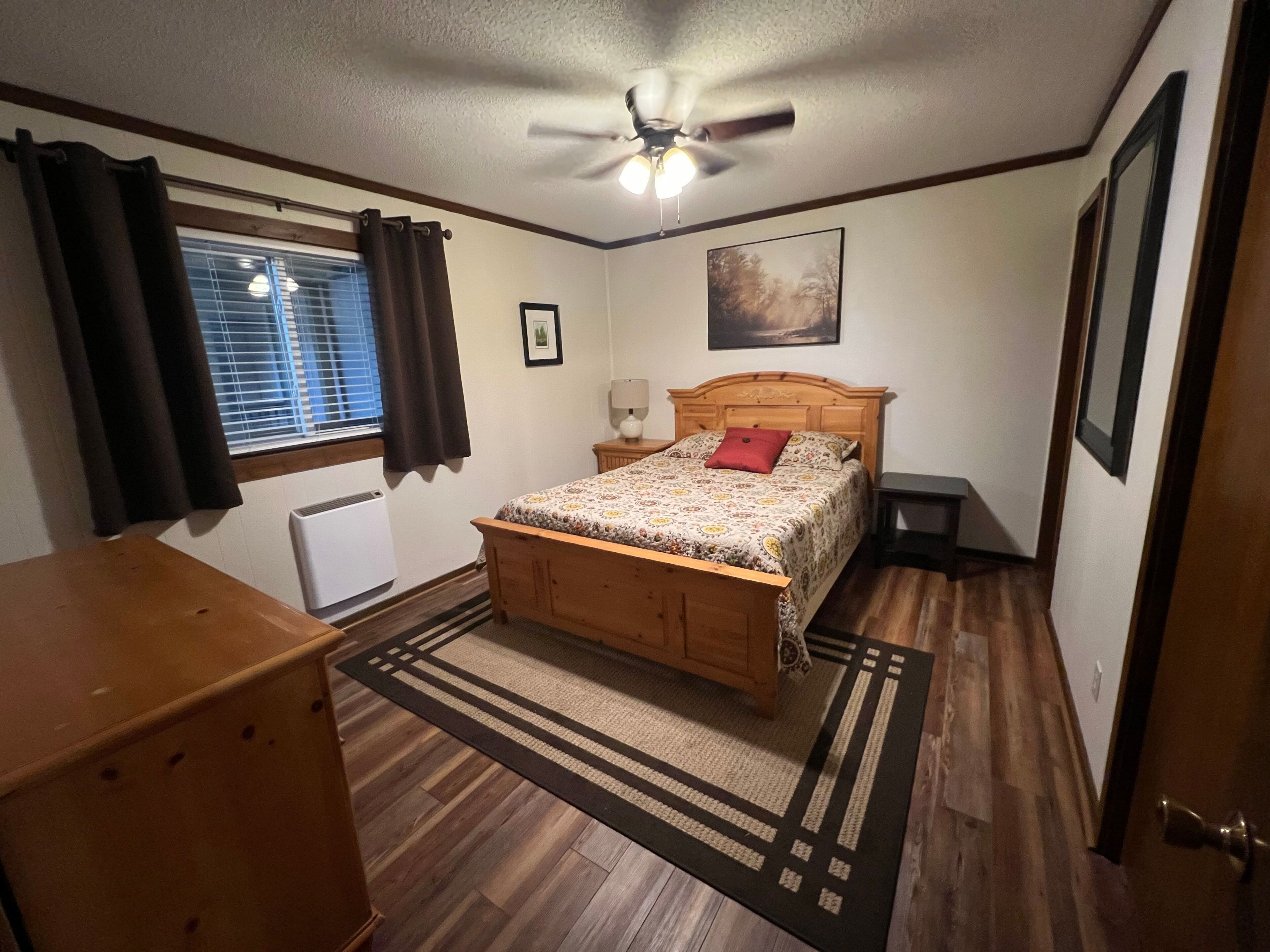 A bedroom with a wooden bed, a ceiling fan, a window with curtains, a radiator, and a rug.