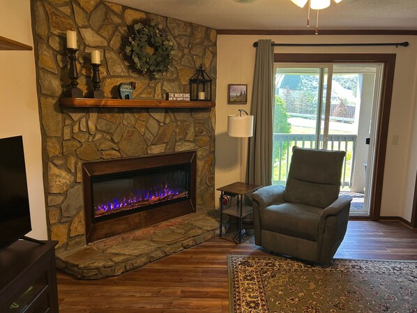 A living room with a stone fireplace, a television, a comfortable armchair, and a view through a glass door.