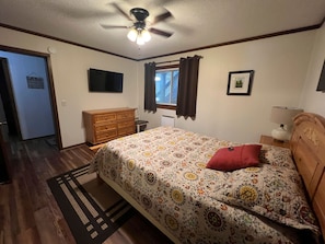 Een slaapkamer met een bed, een dressoir, een televisie, een raam met gordijnen en een plafondventilator.