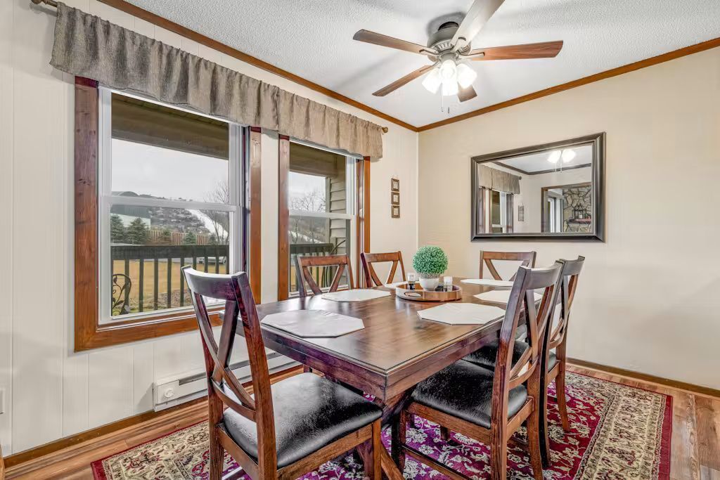 A dining room with a wooden table, six chairs, a ceiling fan, and large windows.