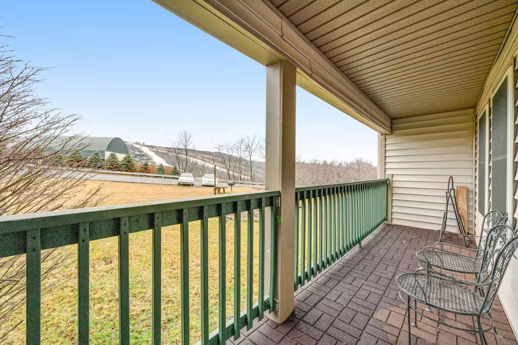 A covered porch with a green railing, outdoor seating, and a view of a field and distant buildings.
