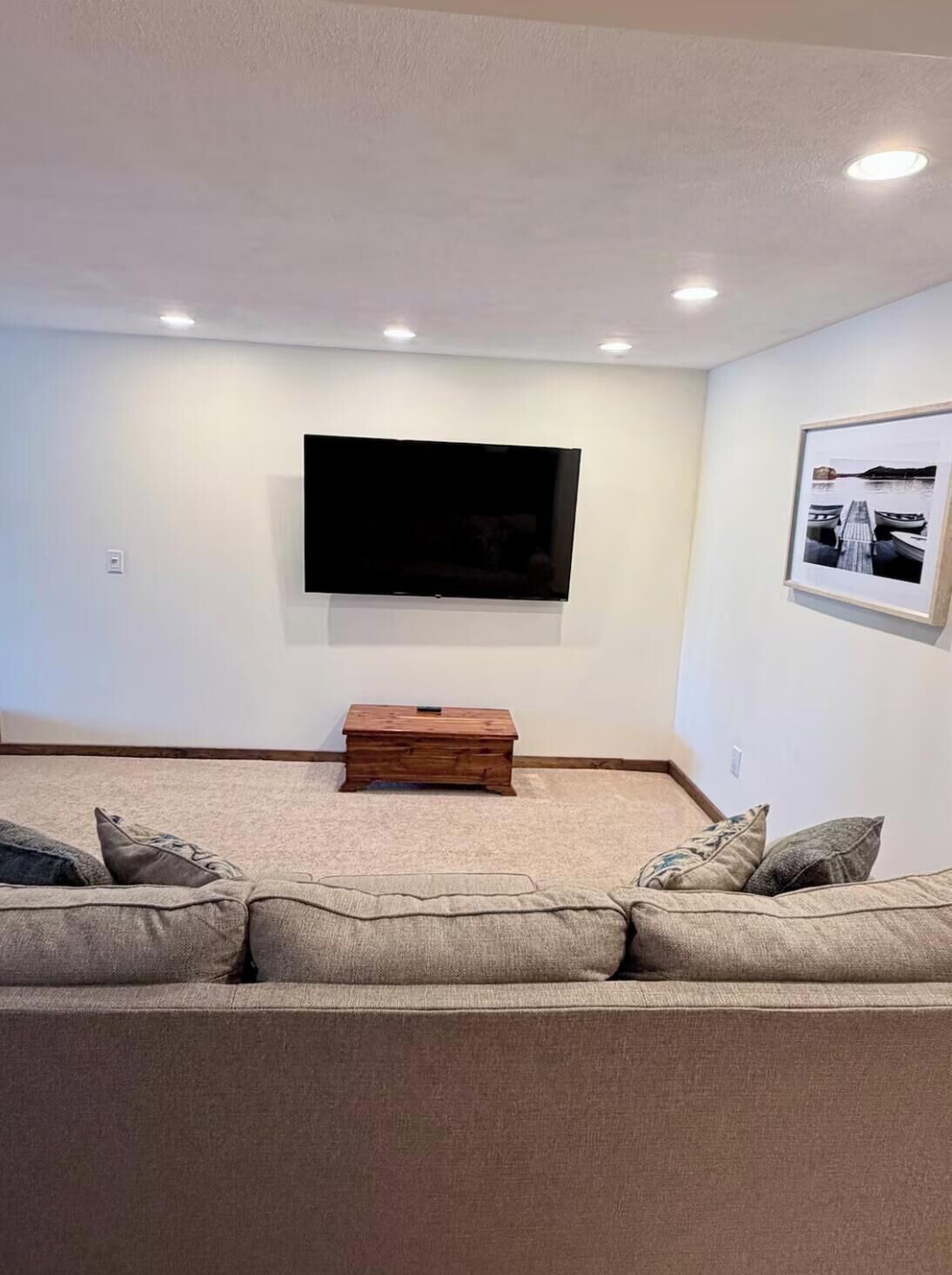 A living room with a flat-screen TV mounted on the wall, a wooden coffee table, and a beige sofa with cushions.