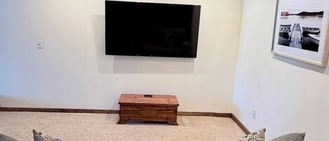 Ein Wohnzimmer mit flachbildschirm-Fernseher an der Wand, einem Holztisch und einem beigefarbenen Sofa mit Kissen.