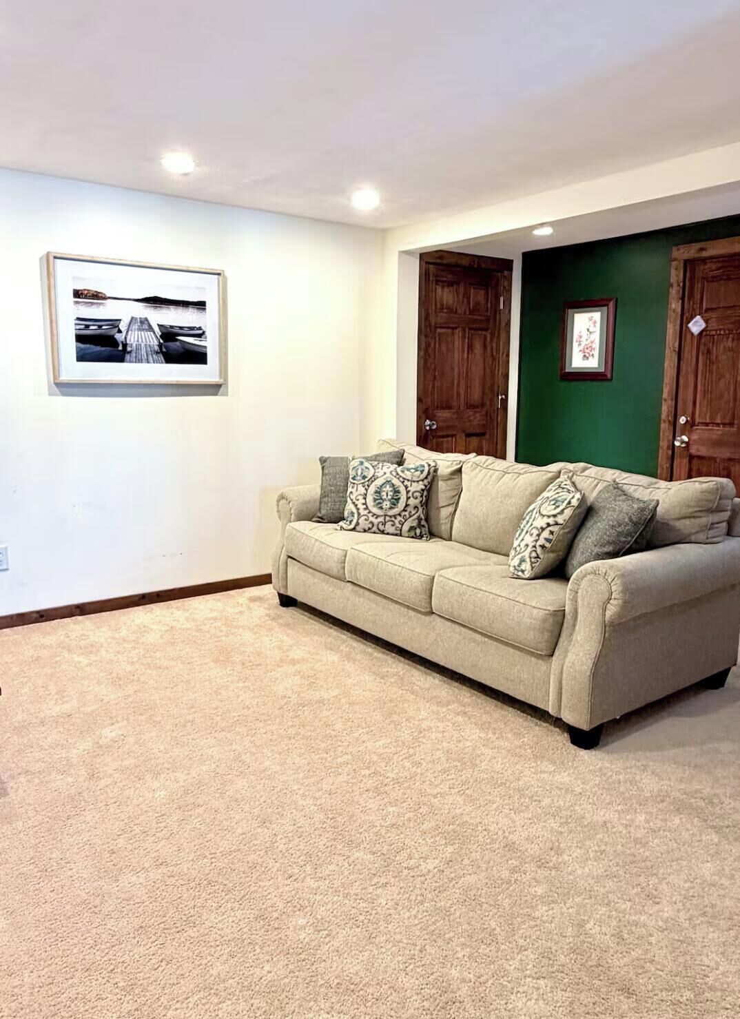 A living room with a beige sofa, patterned cushions, a framed picture on the wall, and a green accent wall.