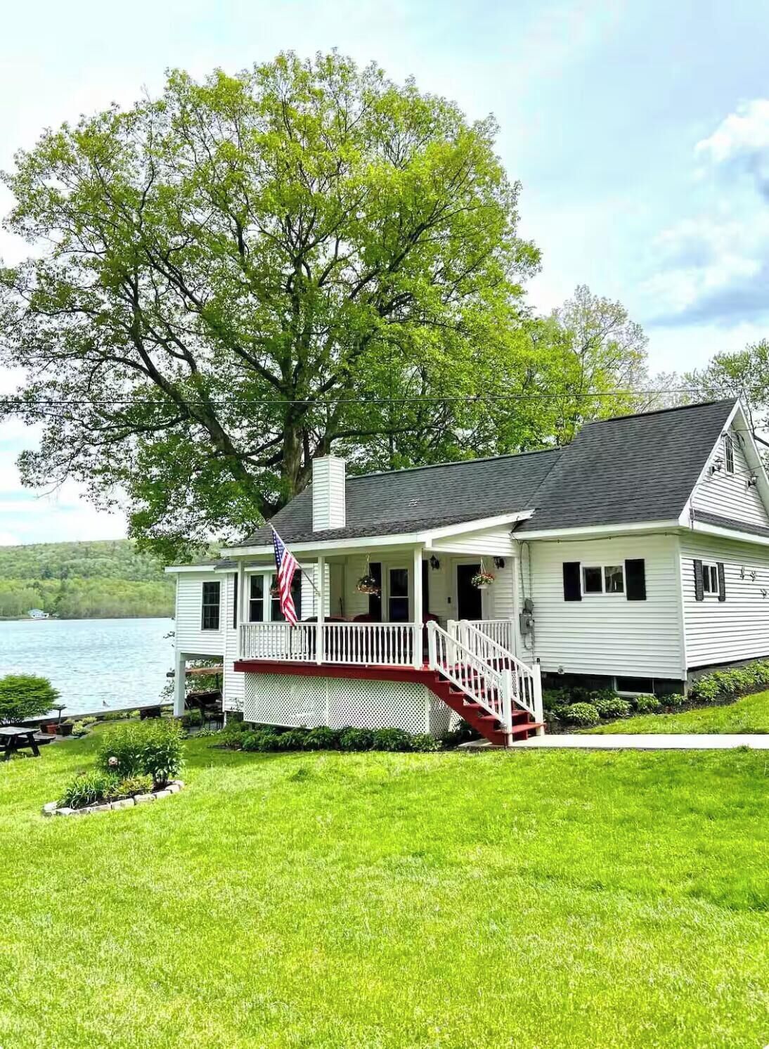 A white house with a porch and a flag, located near a body of water.