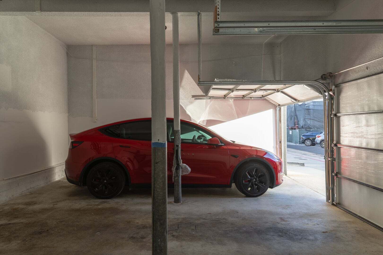 Un auto rojo estacionado dentro de un garaje con puerta metálica y piso de concreto.