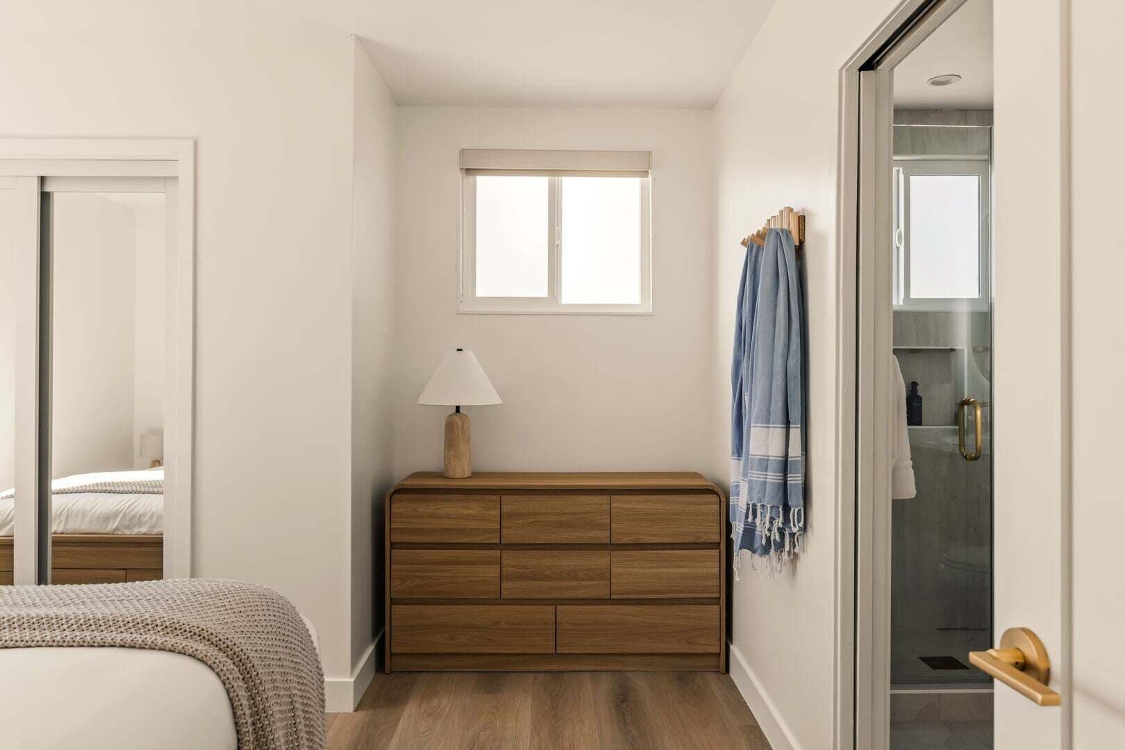 A bedroom with a wooden dresser, a bedside lamp, a window, and a bathroom visible through an open door.
