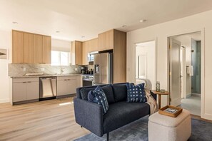 Ein modernes Wohnzimmer mit einer Couch, einem Hocker und einer Küchenzeile mit Schränken und Geräten.
