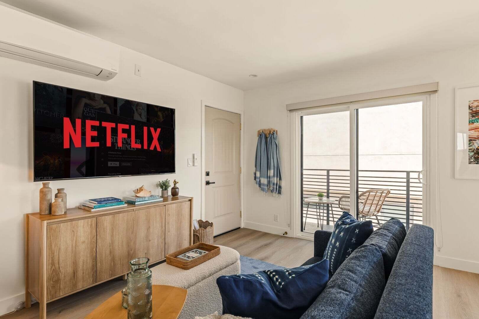 A living room with a large flat-screen TV mounted on the wall, a wooden cabinet, a blue sofa, and a balcony with outdoor furniture.