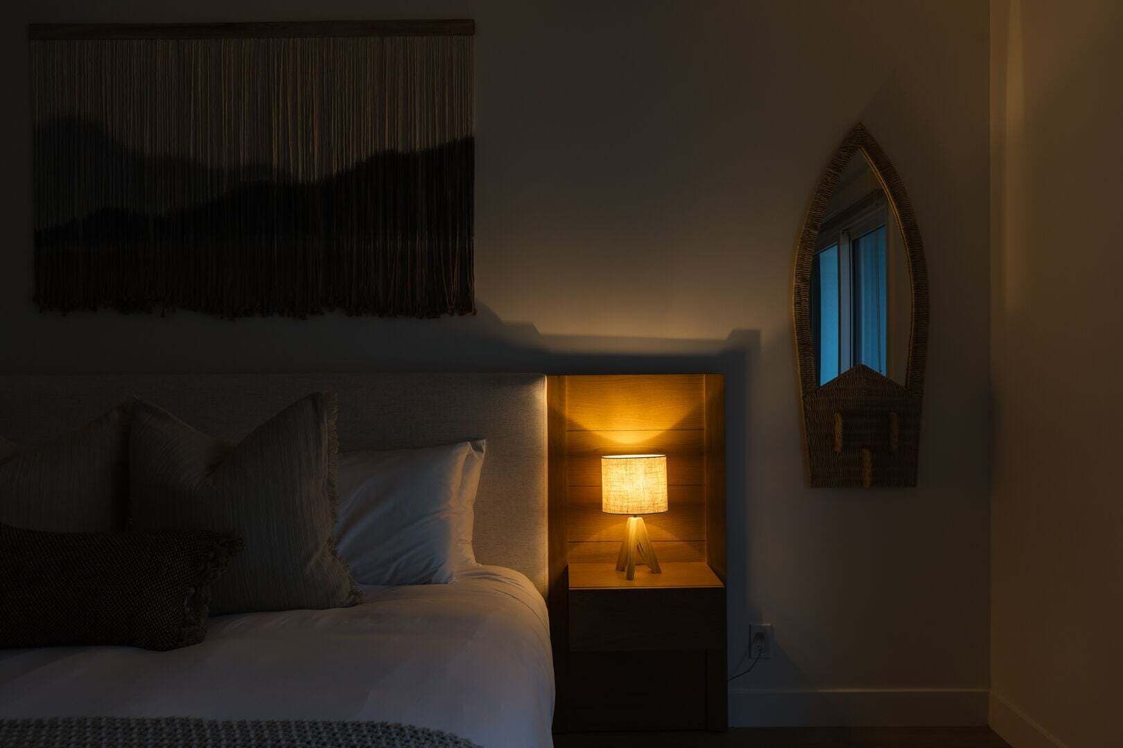 A bedroom with a bed, a nightstand with a lamp, a wall-mounted mirror, and a textured wall hanging.