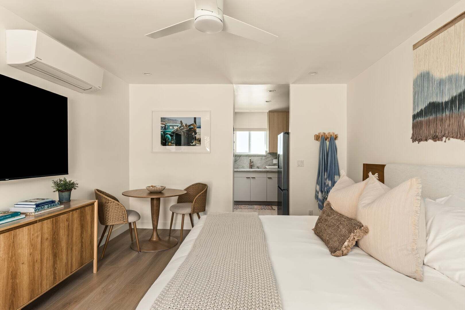 Un dormitorio moderno con una cama grande, un escritorio, una silla, un televisor y una cocina al fondo.