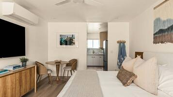 Un dormitorio moderno con una cama grande, un escritorio, una silla, un televisor y una cocina al fondo.