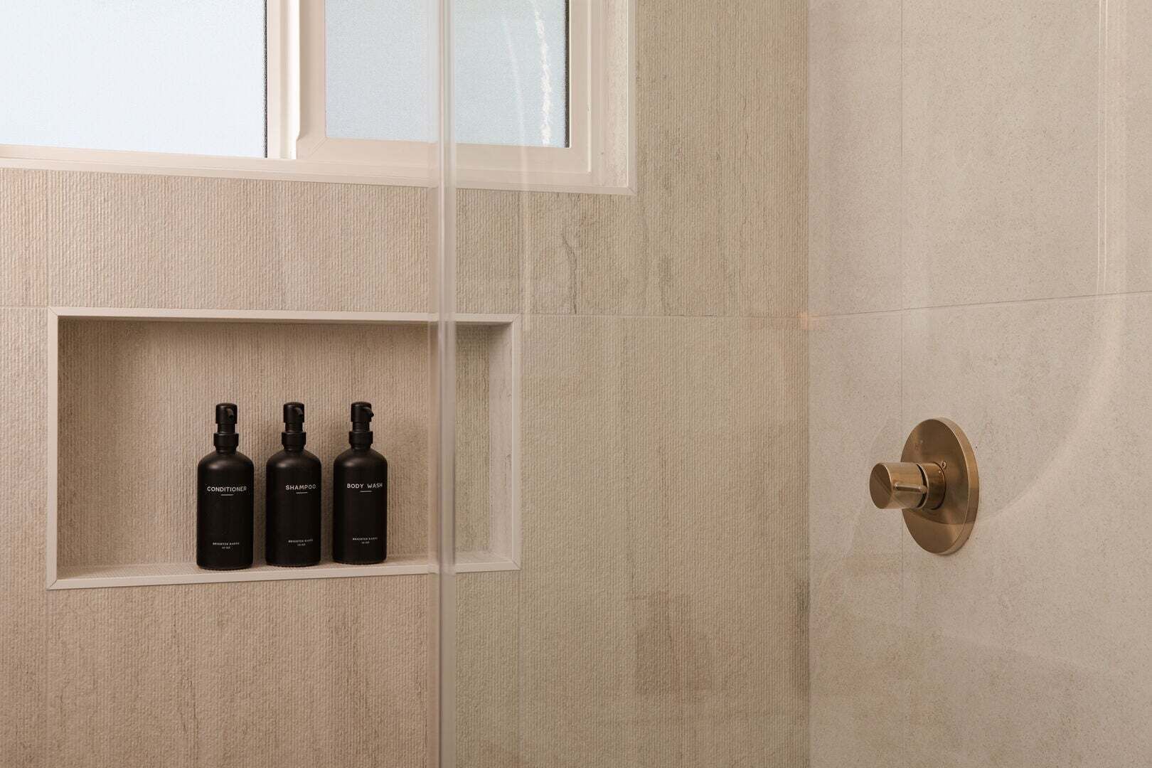 A bathroom with a marble wall, a window, and a shelf with three bottles labeled 'CONDITIONER,' 'SHAMPOO,' and 'BODY WASH.'