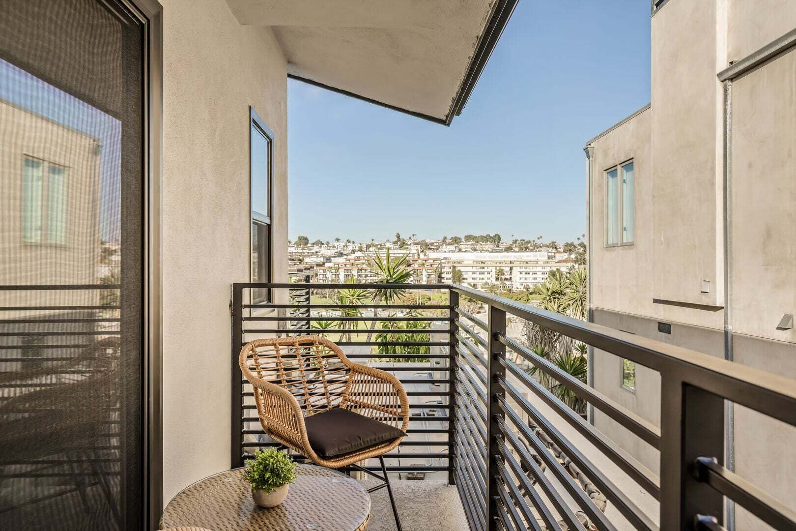 Ein Balkon mit einem Korbstuhl und einem kleinen Tisch mit einer Pflanze, mit Blick auf eine Wohngegend.