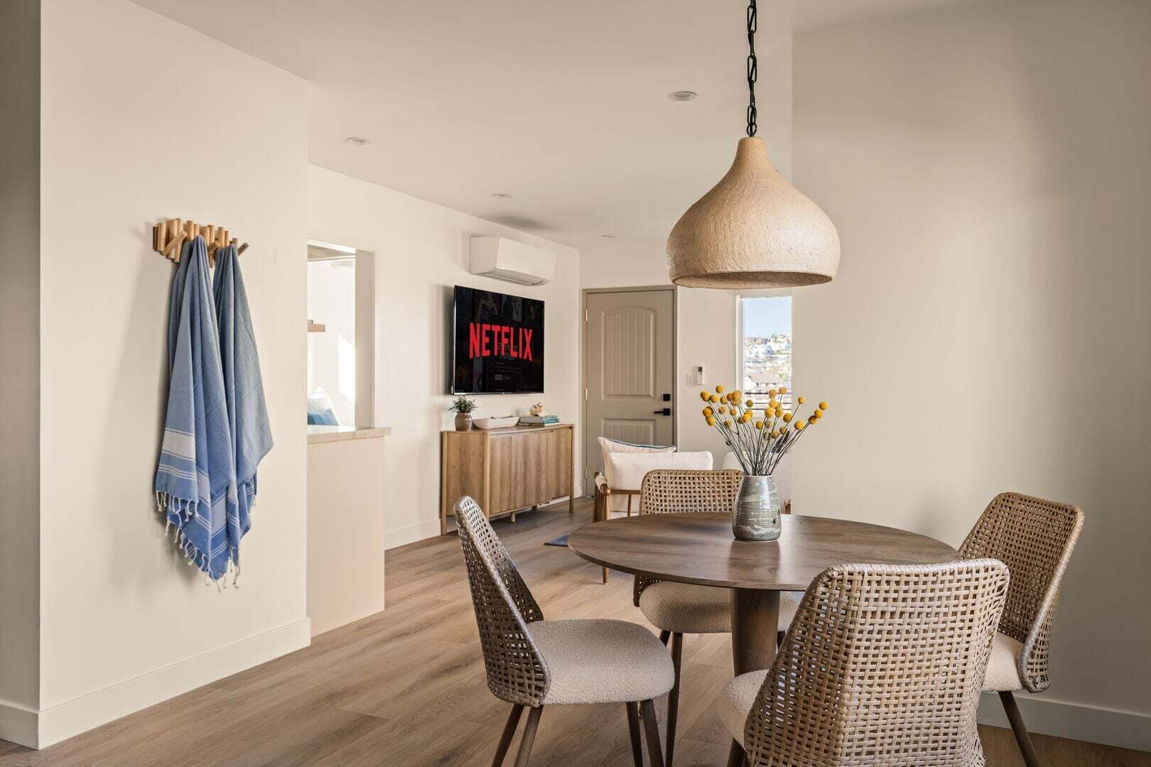 Un coin repas avec une table ronde, des chaises en osier, un luminaire suspendu et un téléviseur fixé au mur qui affiche « NETFLIX ».