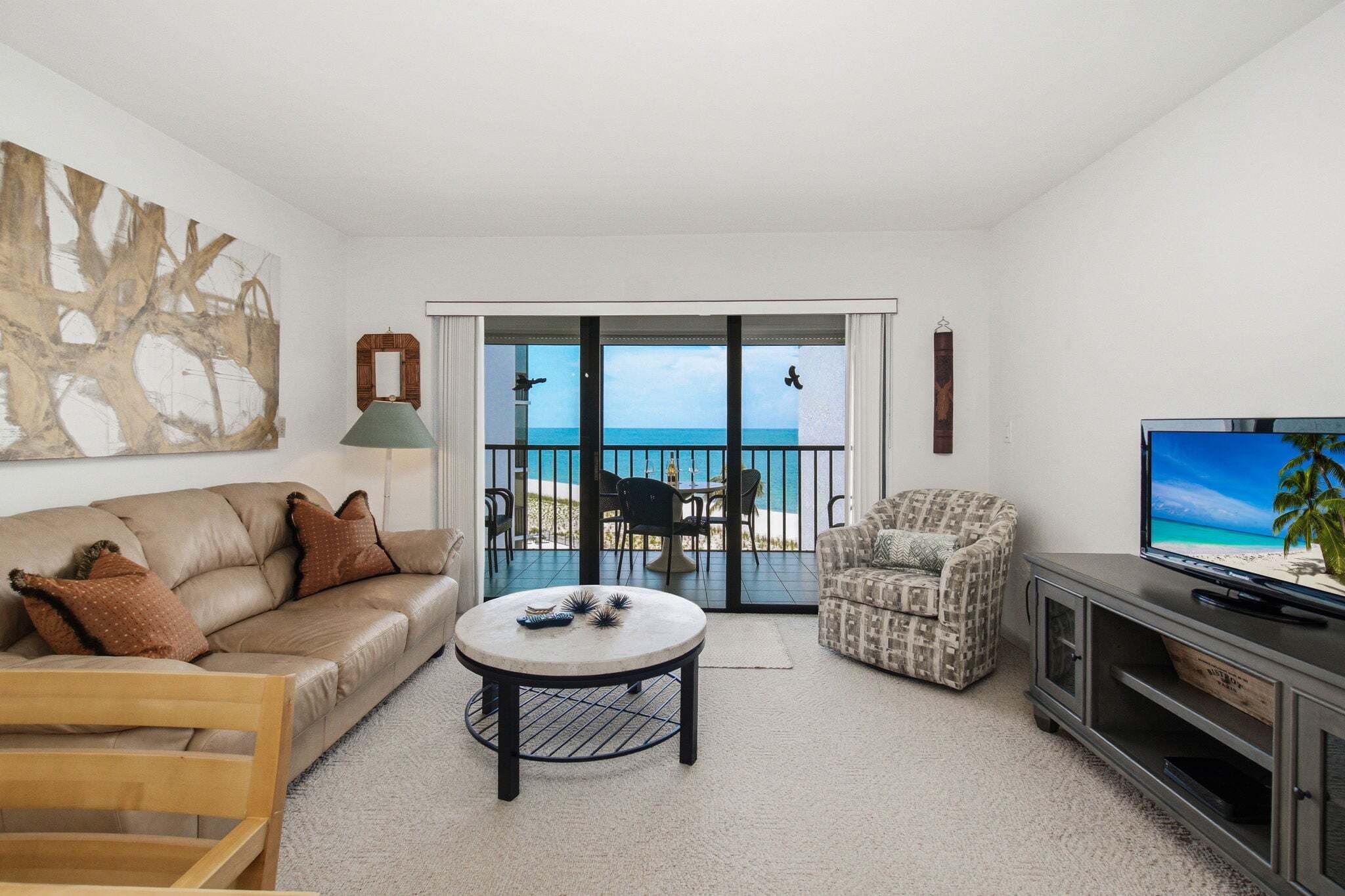A living room with a sofa, a round coffee table, a patterned armchair, a television, and a balcony with a view of the sea.