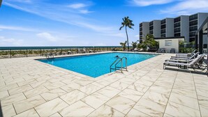 A swimming pool with a clear view of the ocean, surrounded by lounge chairs and a building with a modern design. - Beachfront 2BR Condo with Gulf Views, Screened Lanai & Pickleball | Sleeps 4 (Bonita Springs)
