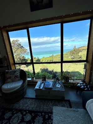 A room with a large window offering a view of the sea and hills. There is a sofa, a small table with a book and remote control, and a rug.