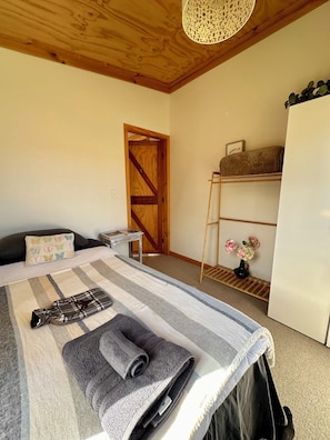 A bedroom with a bed, a wooden door, a small table, a shelf with flowers, and a white refrigerator. - Bang for your buck, Sanctuary, Sea & Starry nights, 5 minutes from Gore Bay! (Domett)