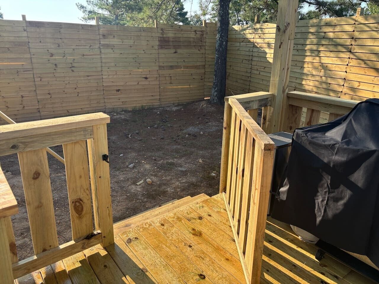 Una terraza de madera con zona de parrilla cubierta, rodeada por un cerco de madera y un árbol.