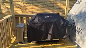 A barbecue grill with a black cover, a trash can, and a wooden deck.