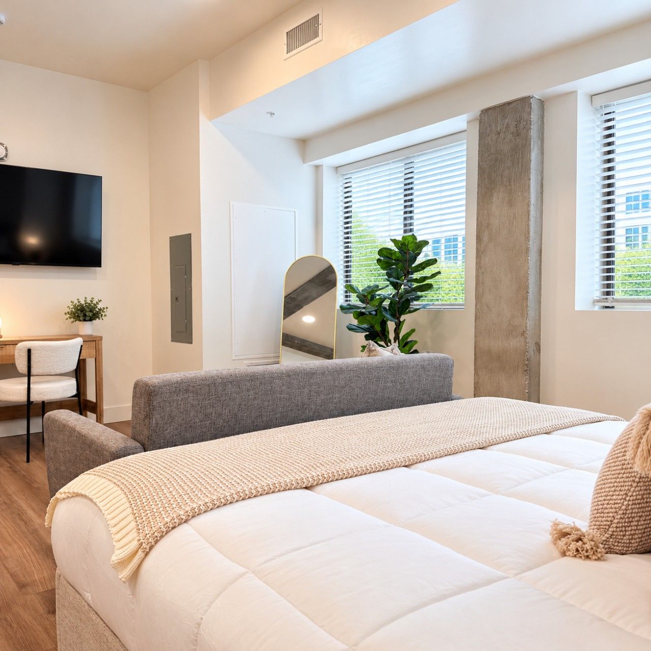 A modern bedroom with a large bed, a flat-screen TV mounted on the wall, a desk with a chair, and a window with blinds.