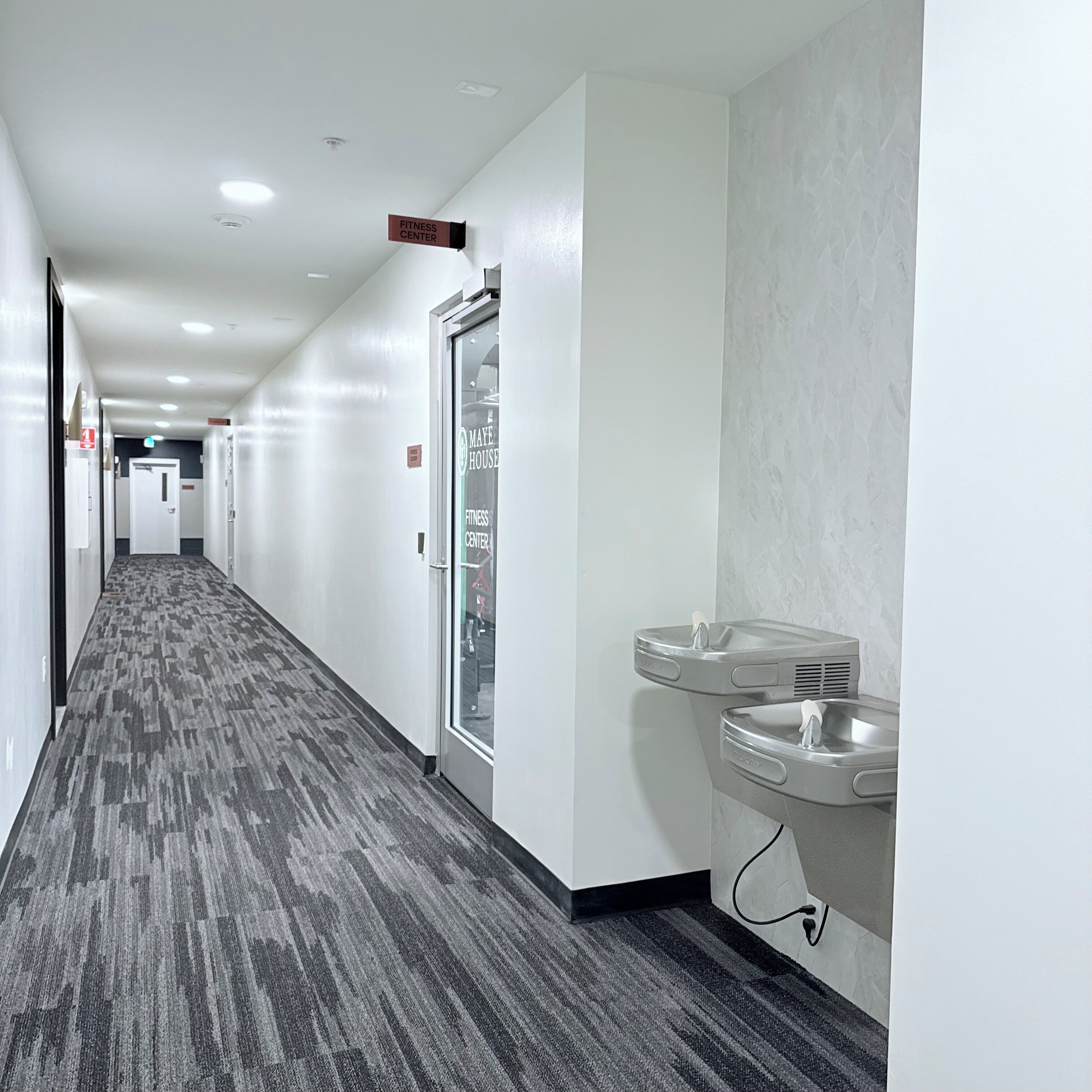 Ein Hotelgang mit einem Fitnesscenter-Schild, einer Glastür und einem Waschbecken mit Handtuchspender.