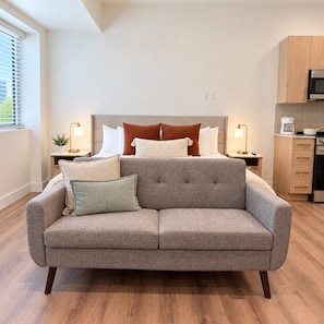 A modern bedroom with a grey sofa, a bed with multiple pillows, bedside tables with lamps, and a kitchenette area. - Downtown SLC Studio | King Bed + Free Parking (Salt Lake City)