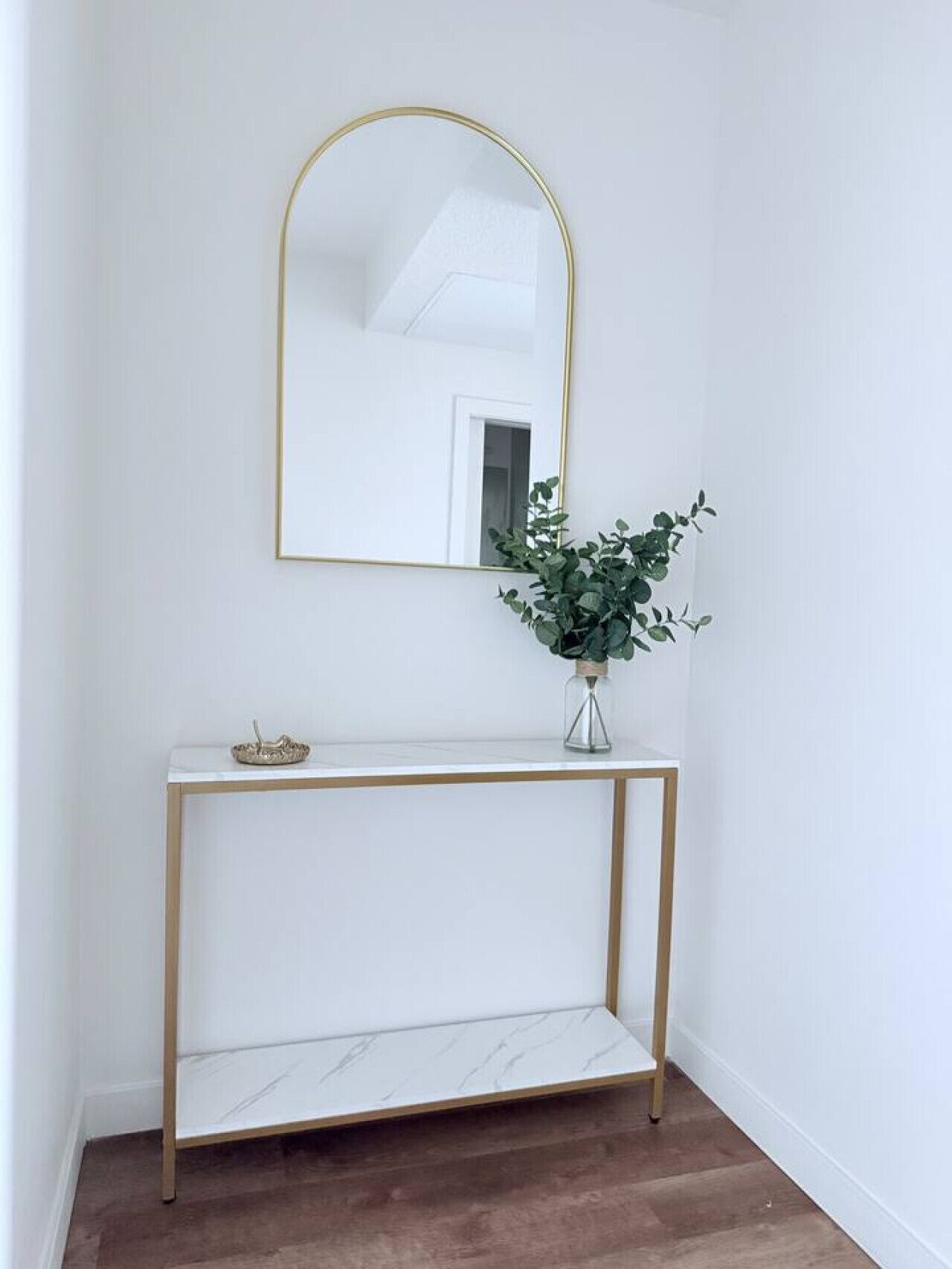 Une console blanche avec un cadre doré, un petit vase contenant des feuilles vertes et un miroir placé au-dessus.