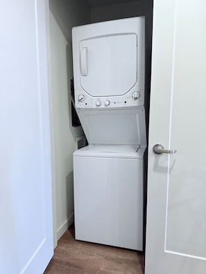 A compact, white laundry room with a front-loading washer and dryer, a door handle, and a small window. - Downtown SLC Studio | King Bed + Work Space (Salt Lake City)