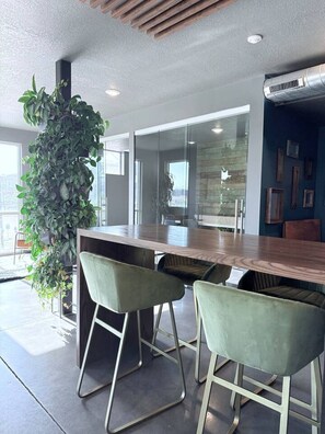A modern office space with a wooden conference table, two green bar stools, a large potted plant, and a glass partition. - Downtown SLC Studio | King Bed + Work Space (Salt Lake City)