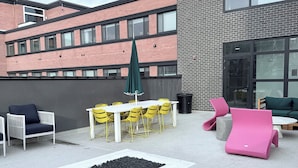 Et udendørs opholdsområde med borde, stole, en bålplads og moderne møbler foran en bygning.