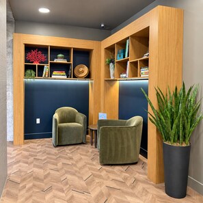 Ein Zimmer mit Fischgrätparkett, zwei grüne Sessel, eine hohe Topfpflanze und ein Holzregal mit verschiedenen Gegenständen.