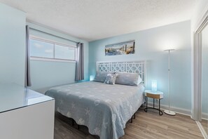 A bedroom with a bed, a bedside table with a lamp, a window with blinds, and a painting on the wall. - 0704 Beach Life-Free Activities Included (Carolina Beach)