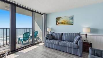 A room with a sofa, two chairs, a painting, and a view of the beach.