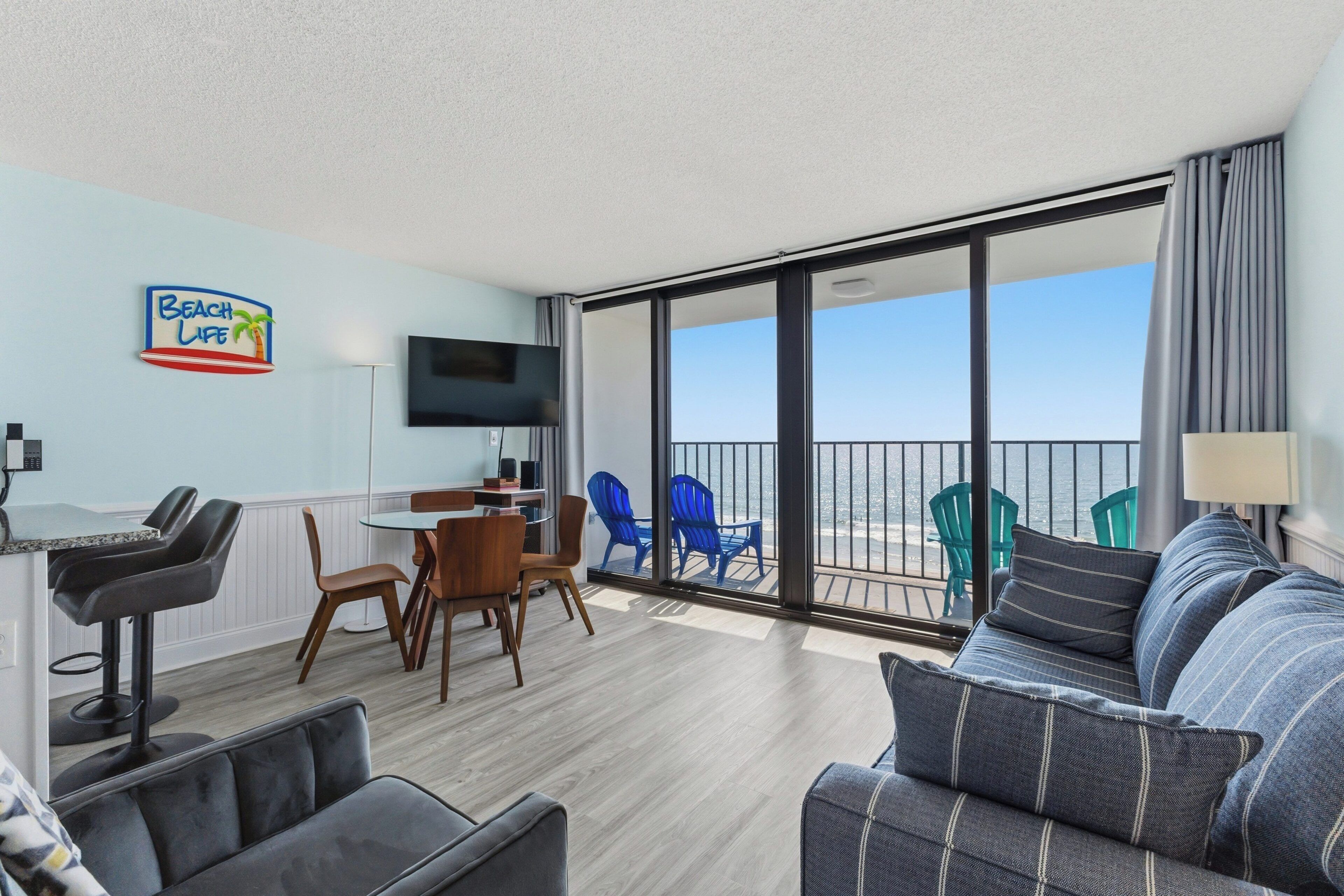 A modern living room with a sofa, chairs, a dining table, and a TV. There is a balcony with a view of the ocean.