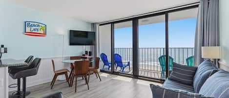 A modern living room with a sofa, chairs, a dining table, and a TV. There is a balcony with a view of the ocean.