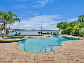 A swimming pool with a curved edge, surrounded by a brick patio, palm trees, and a view of the water. - The Bayfront - Bayfront Pool Spa by Anna Maria Life Vacation Rentals (Bradenton Beach)