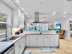 A modern kitchen with white cabinetry, a central island, and a view to the living area. - The Bayfront - Bayfront Pool Spa by Anna Maria Life Vacation Rentals (Bradenton Beach)