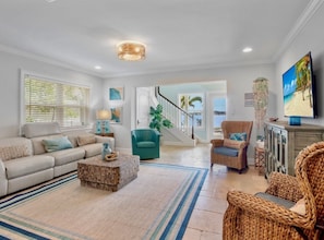 A spacious living room with a sofa, coffee table, and wicker chairs. A staircase leads to a balcony with a view of the ocean. - The Bayfront - Bayfront Pool Spa by Anna Maria Life Vacation Rentals (Bradenton Beach)