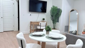 Ein modernes Wohnzimmer mit einem runden Esstisch, weißen Stühlen, einem flachbildfernseher an der Wand, einem großen Spiegel und einem Blumentopf.