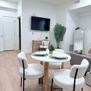 Una sala de estar moderna con una mesa de comedor redonda, sillas blancas, un televisor de pantalla plana montado en la pared, un espejo grande y una planta en maceta.