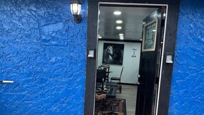 Pared exterior azul con base de piedra, una puerta de cristal que da a un espacio interior con zona de comedor y una lámpara fijada a la pared.