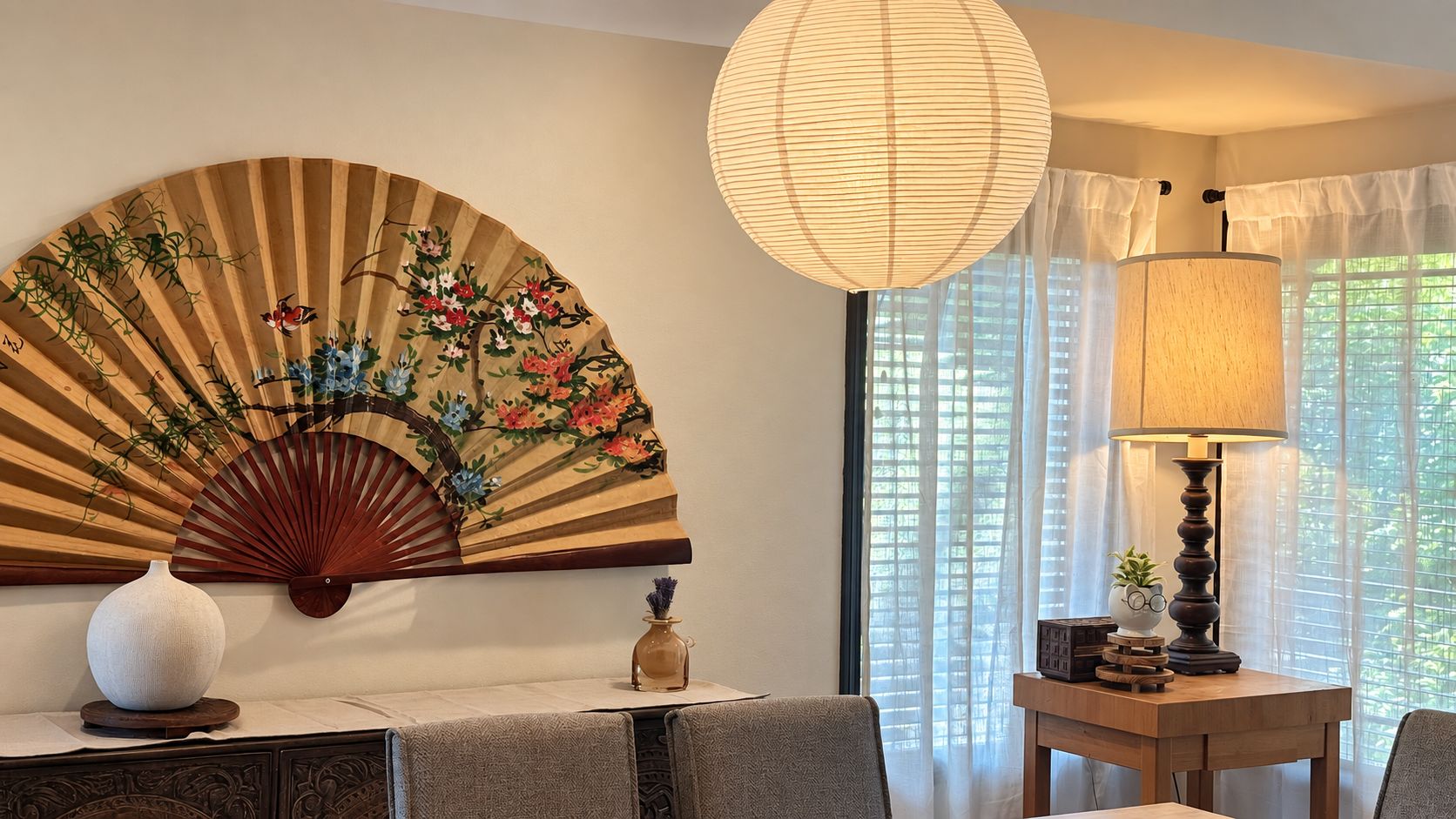 A room with a traditional fan, a table with a lamp, and a view through sheer curtains.