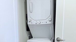 A compact, white laundry room with a front-loading washer and dryer, a door handle, and a small window. - Modern Downtown SLC | King Bed + Free Parking (Salt Lake City)